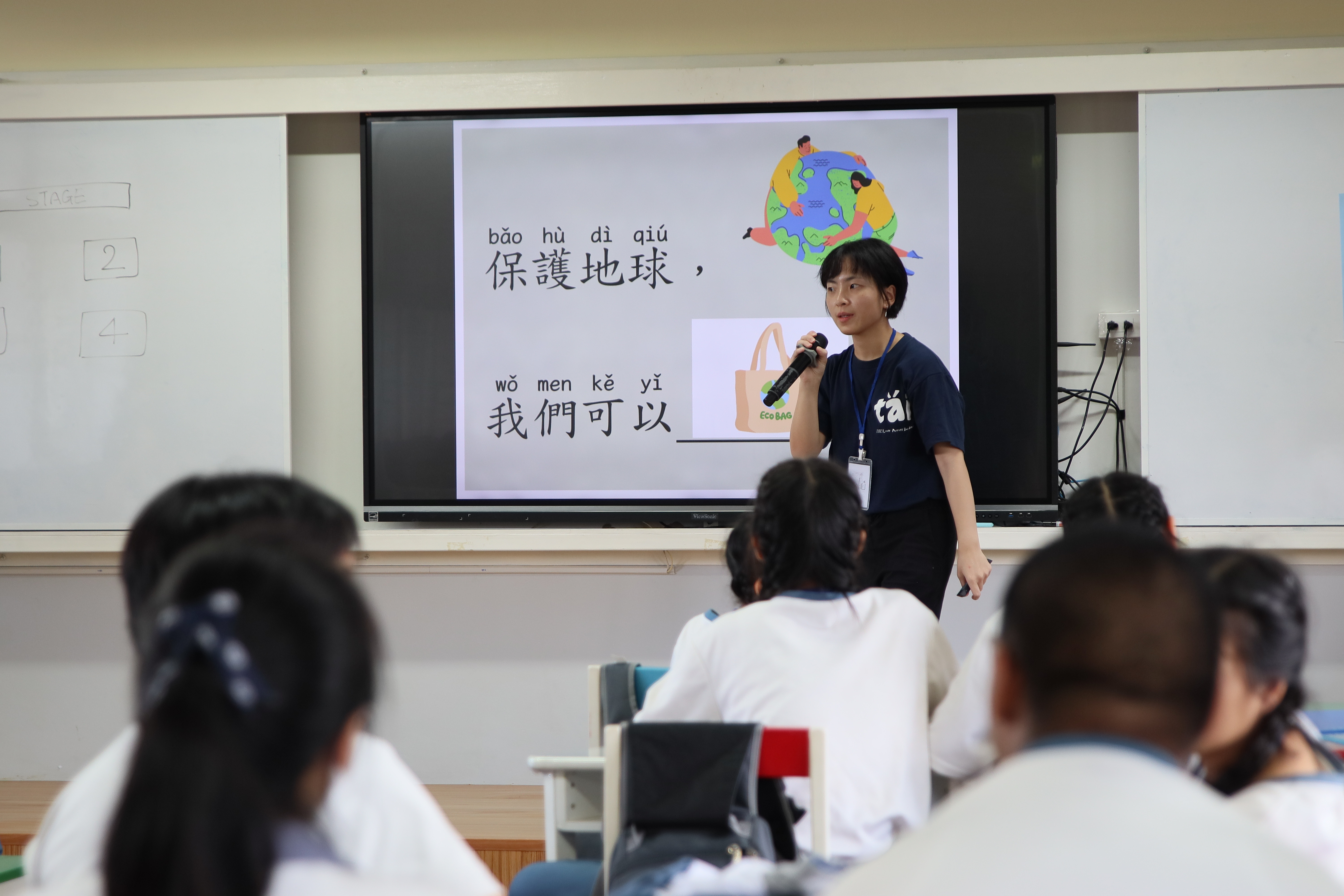 志工老師在教室前方以簡報方式，向學生們介紹「保護地球」的主題，並以華語及拼音教學，鼓勵大家一起響應環保行動。學生們認真聆聽，展現積極學習態度。